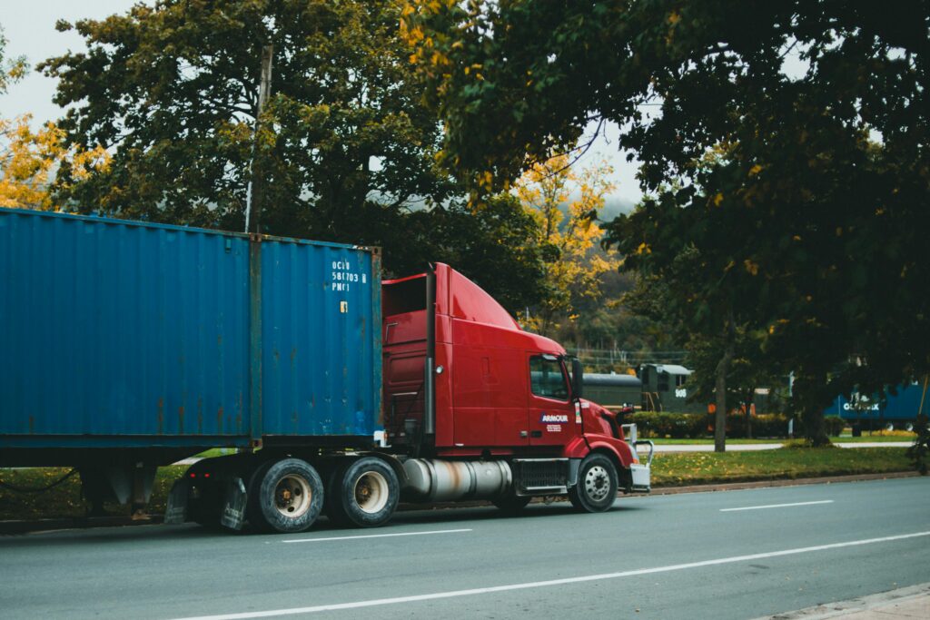A red truck transports a blue cargo container on a scenic, tree-lined road.